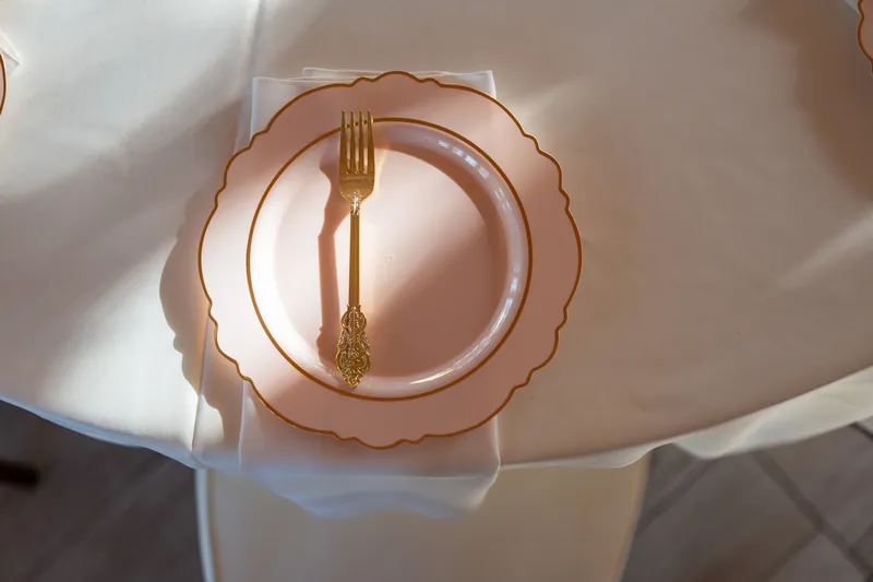 A rose-gold scalloped plate and matching gold fork rest on white linens, with sunlight casting shadows across the tablescape.