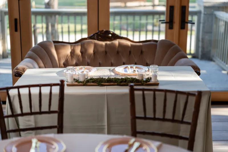 A taupe velvet settee frames the sweetheart table decorated with greenery along the railing overlooking the venue grounds.