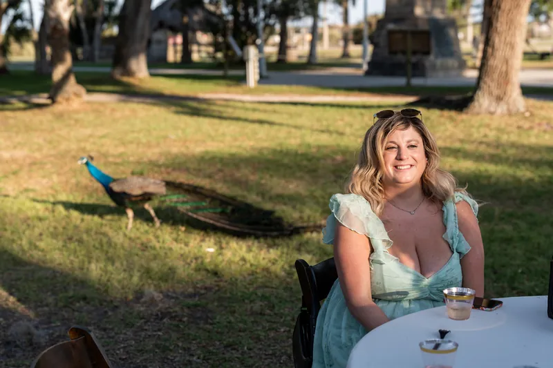 Lena smiles while seated at an outdoor table with a peacock perched nearby at Fountain of Youth Archaeological Park.