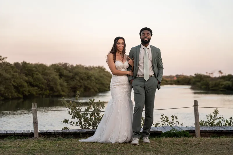 Lena and Blake pose together by the water's edge at dusk, with Lena in her flowing white gown and Blake in a gray suit.