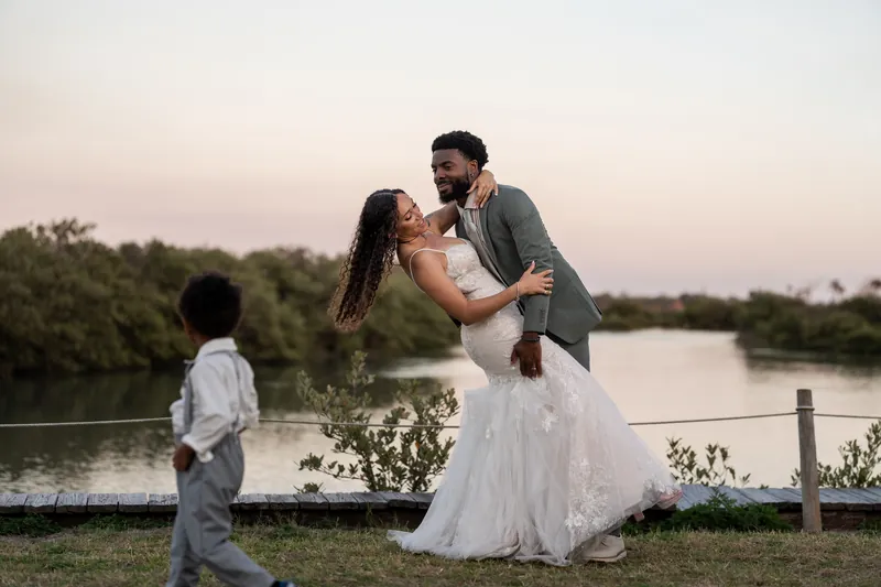 Blake lifts Lena in a kiss while their son watches from the grass beside them near the water.