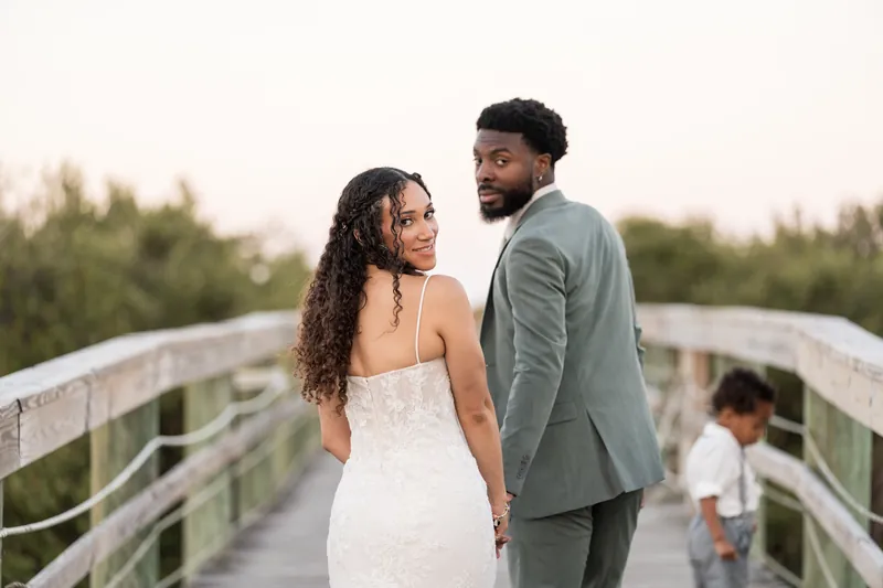 Lena in a strapless white wedding dress and Blake in a gray suit stand face-to-face on a wooden boardwalk at Fountain
