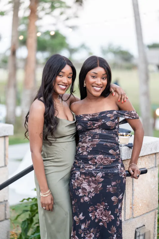 Two women in sage green and floral dresses smile warmly at the camera with trees behind them.