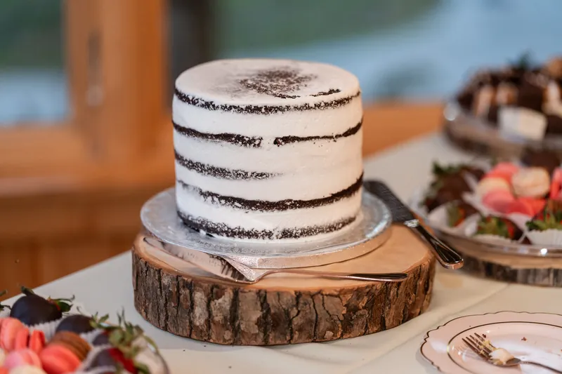 A tiered white cake with dark chocolate bands sits on a wood-slice stand surrounded by floral decorations.