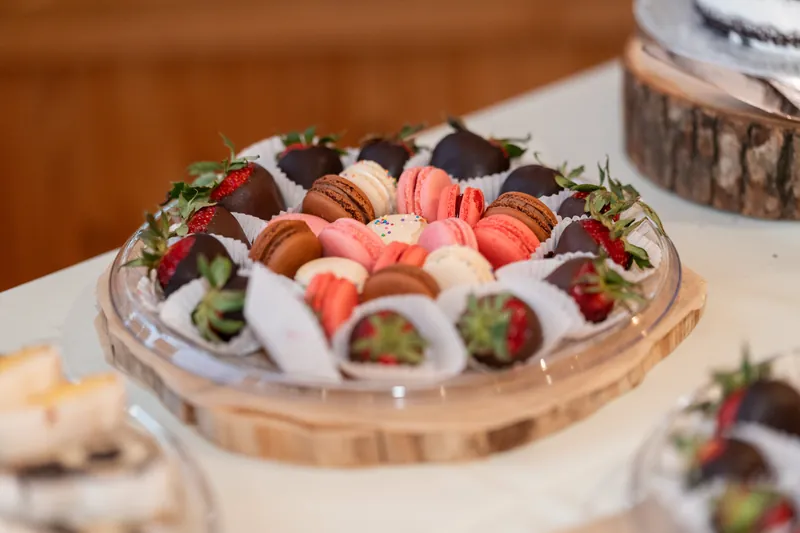 Chocolate-covered strawberries and macarons arranged on a wooden platter with pink flowers nearby.