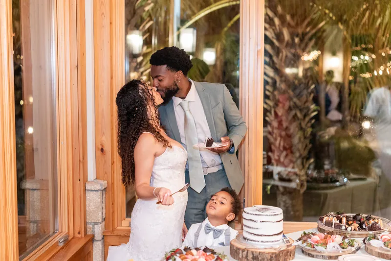 Lena and Blake share a kiss while their young son stands between them at the reception, with desserts displayed on wood-slice