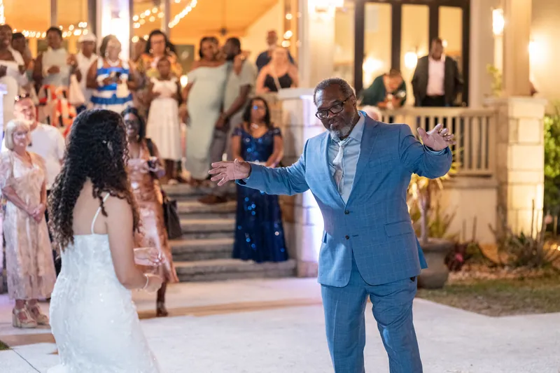 Lena dances with a guest in a blue suit while smiling broadly, with other guests and the lit venue visible behind them.