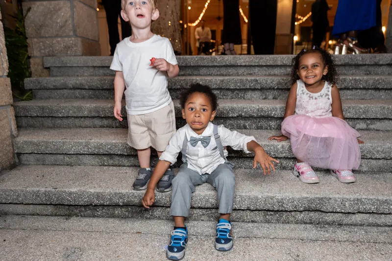 Three young children sit on stone steps with the glowing reception pavilion and string lights visible behind them.
