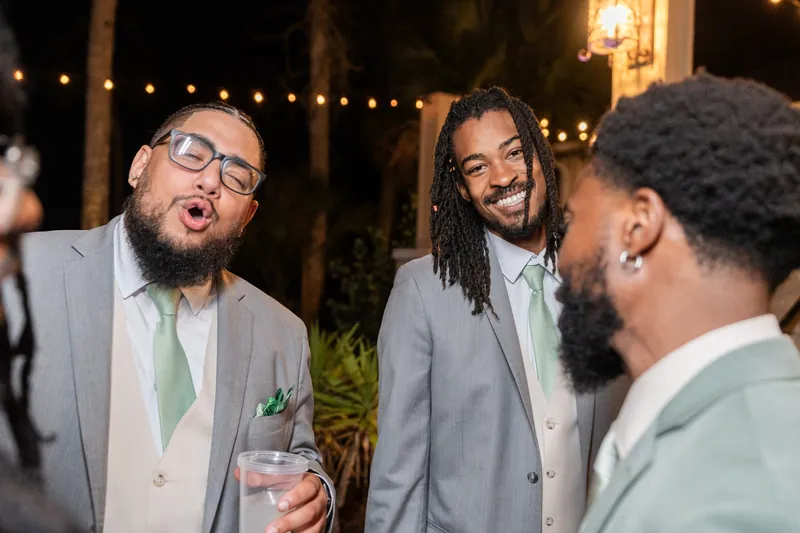 Three men in suits laugh and chat under string lights and palm trees at Fountain of Youth Archaeological Park, St. Augustine.