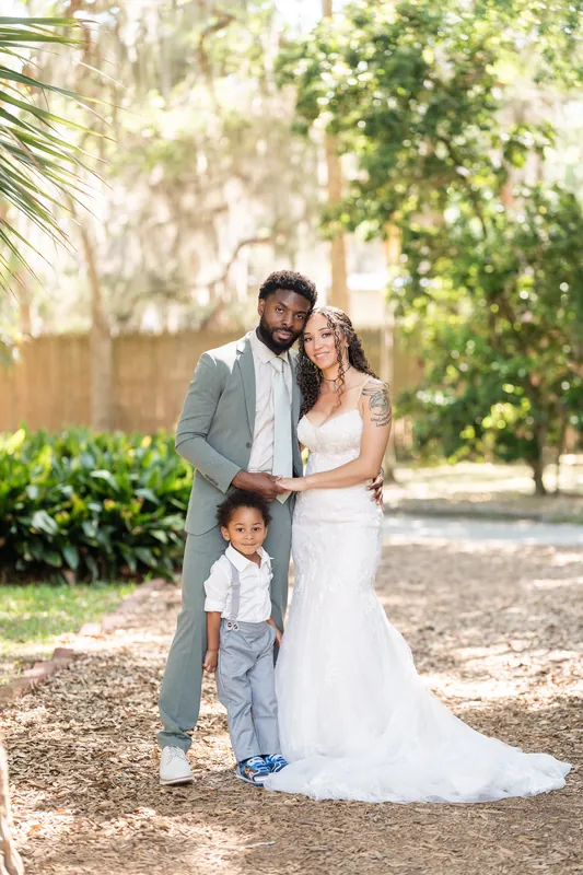 Blake and Lena pose with their young son between them on a tree-covered lane, all three dressed formally.