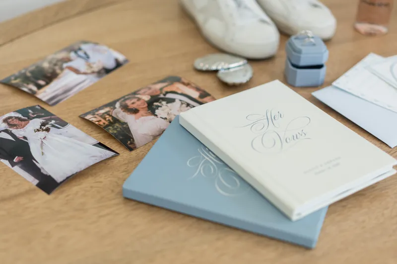 Wedding stationery and photos arranged on a wooden table, including blue and white programs and a pair of white shoes.