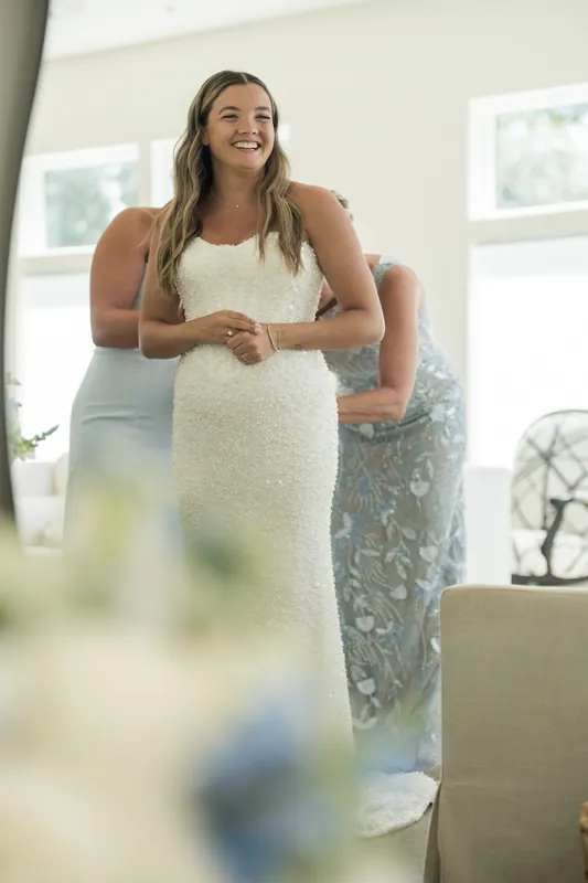 A person in a white wedding dress smiles while adjusting their bouquet, with another person visible behind them in soft focus.