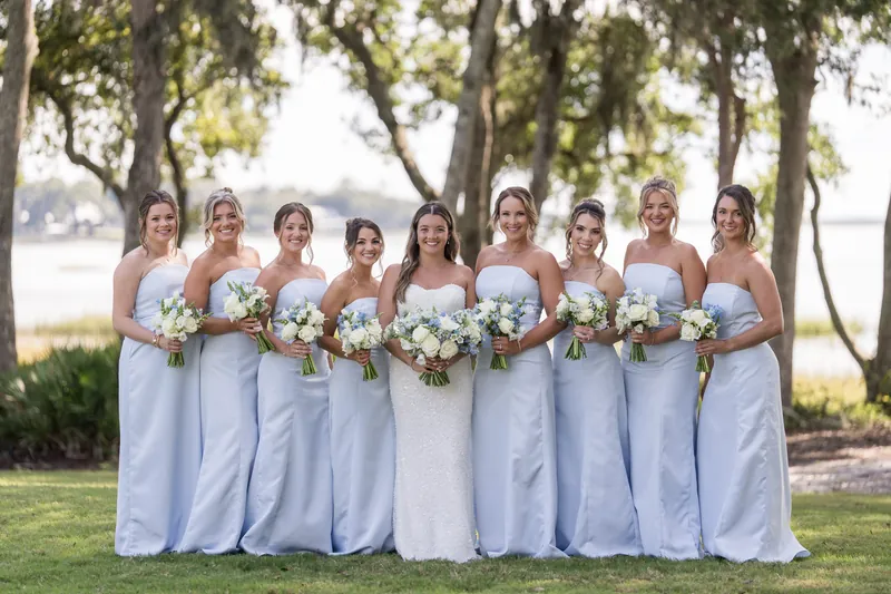 Ten people in lavender and white gowns stand in a line on the lawn, each holding a white and greenery bouquet.