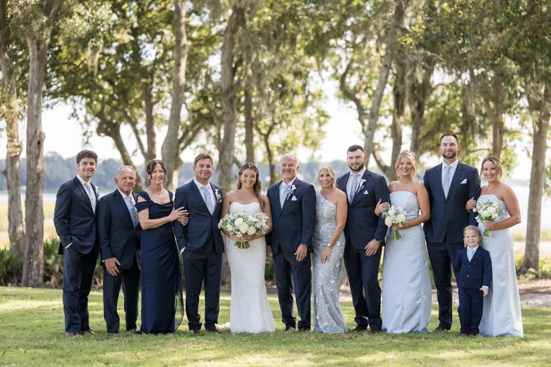 A group of fourteen people in navy suits and white gowns arrange themselves on the lawn for a formal family portrait.