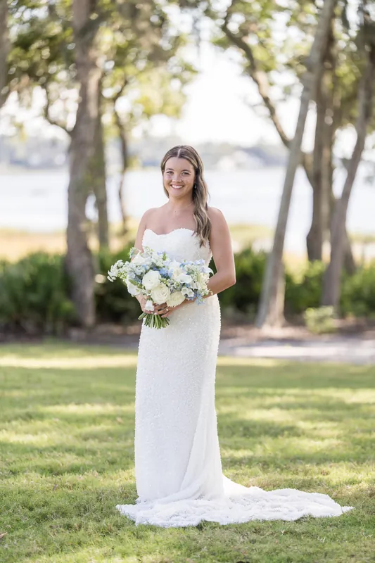 Someone in a flowing white gown stands alone beneath the canopy of oak trees, holding a white and greenery bouquet.
