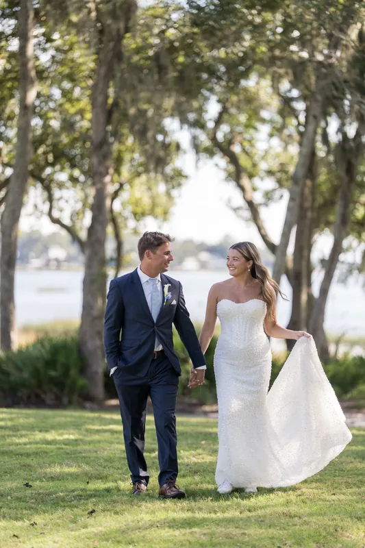 Two people walk across the grass hand in hand beneath the oak trees, trailing a dramatic white gown train.