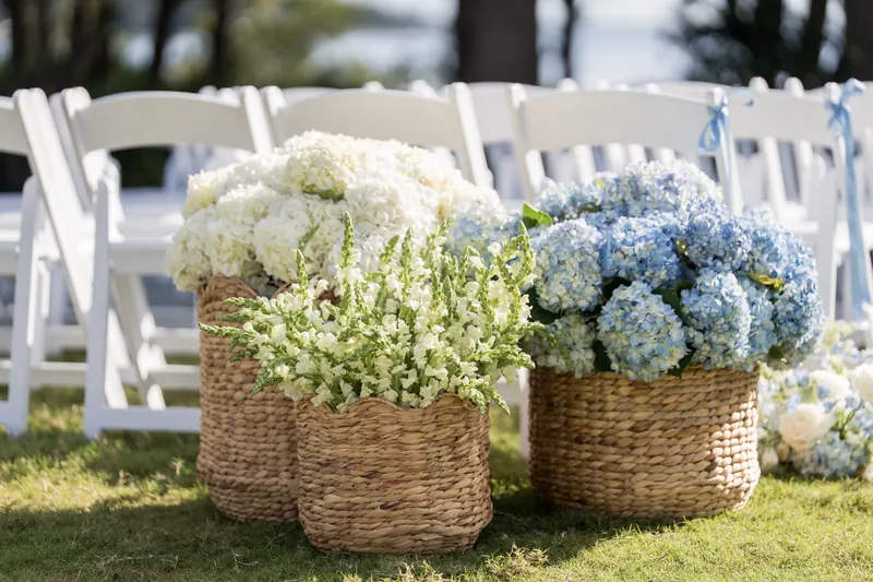 Woven baskets overflow with white hydrangeas and green filler flowers, arranged on grass beside white ceremony chairs.