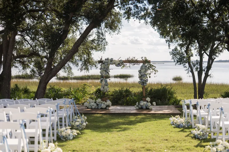 White folding chairs line a riverside ceremony lawn beneath sprawling oaks, with a floral arch visible across the grass.