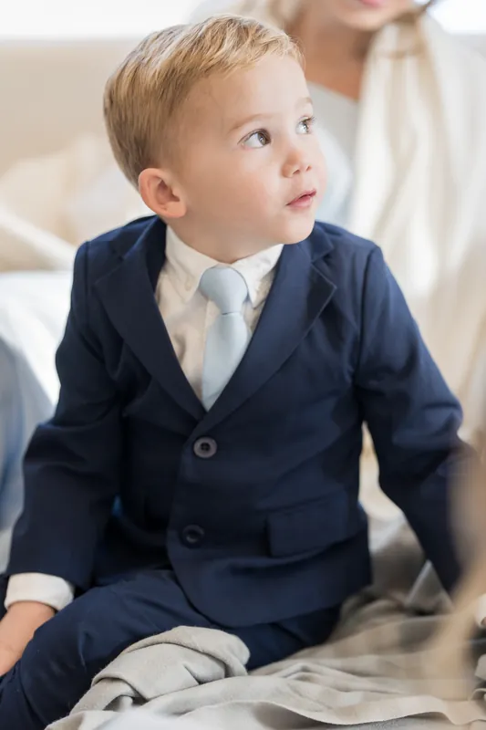 A young child in a navy suit and cream tie sits and gazes upward, dressed for the wedding day.