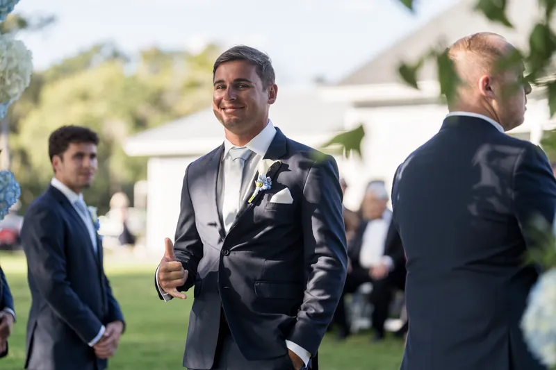 A person in a dark suit stands smiling on the lawn during the reception, with guests mingling in the background.
