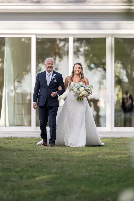A couple walks arm-in-arm across the lawn in front of a white structure, one in a navy suit and one in a flowing white gown.