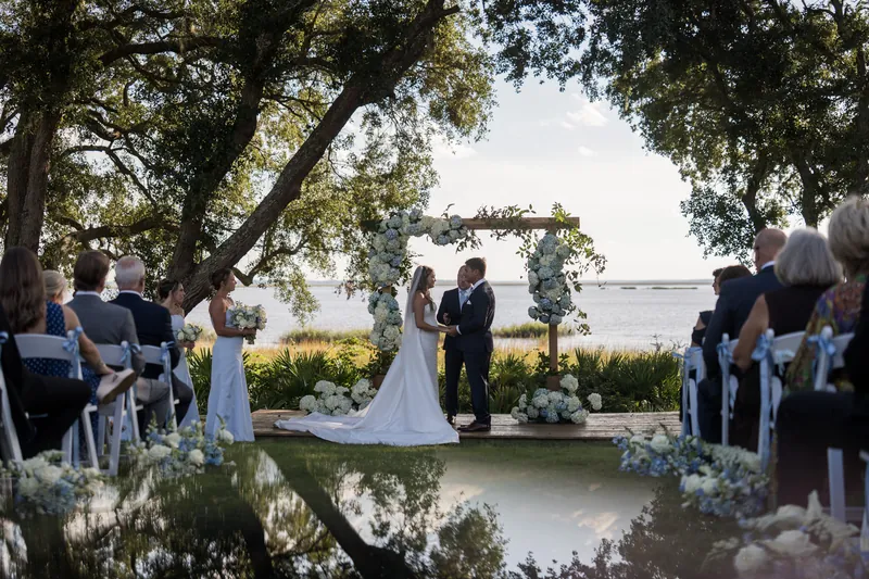 Lexie and Ed stand at an altar over water beneath towering oaks, surrounded by guests in white chairs at The Preserve at Amelia River Club.
