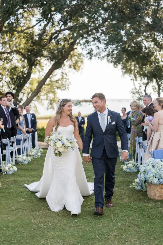 Lexie and Ed walk hand in hand down the grassy aisle after their ceremony, tree-lined and flanked by seated guests.
