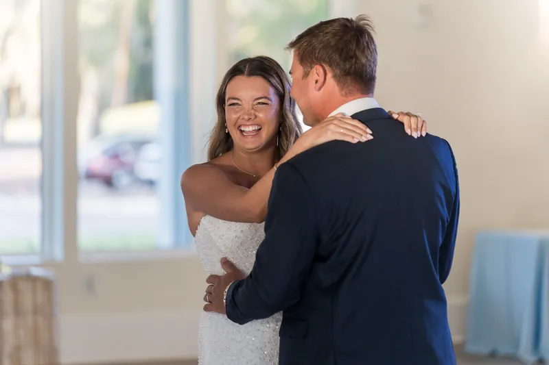 Two people in wedding attire embrace indoors, smiling as natural light streams through nearby windows.
