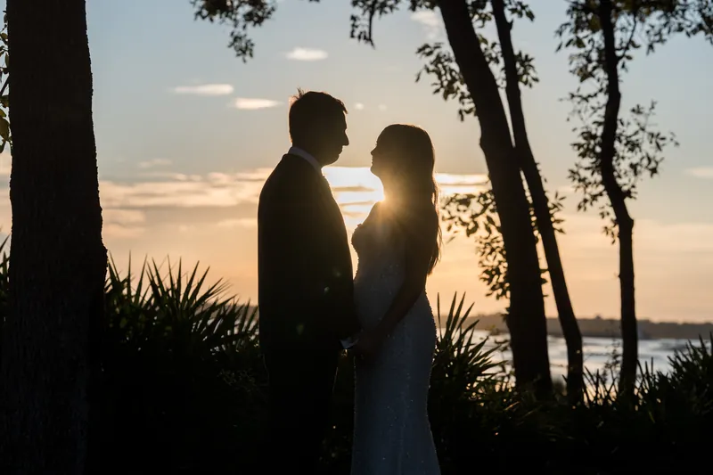 Silhouetted against golden sunset, a couple in formal wear stands close together by the water's edge framed by trees.