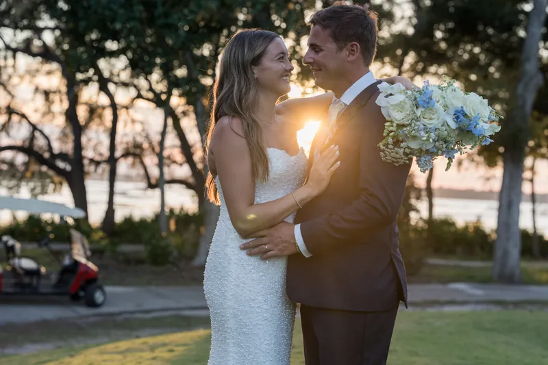 A person in a textured white gown and their partner in a dark suit hold each other as the sun sets behind them near the grounds.