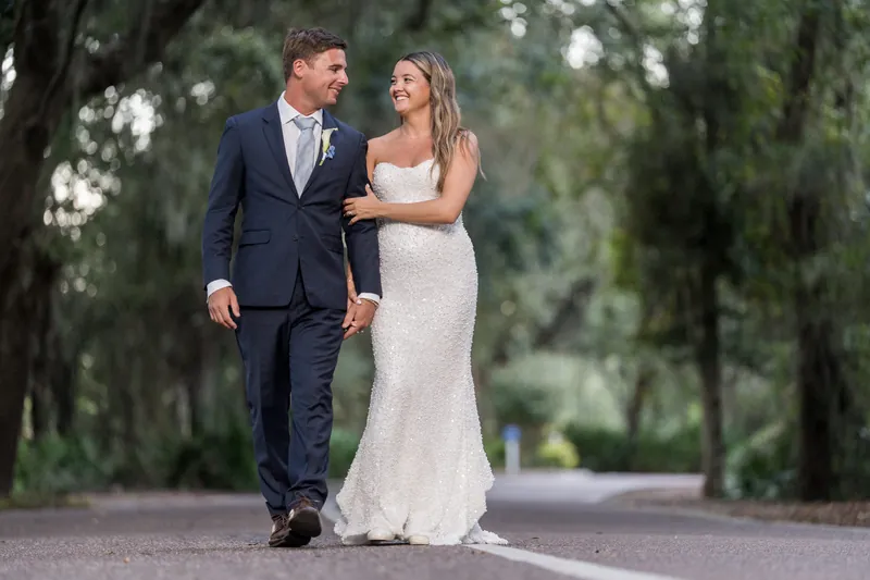Lexie and Ed stroll together down a tree-lined path, her hand resting on his arm as dappled shade falls across them.