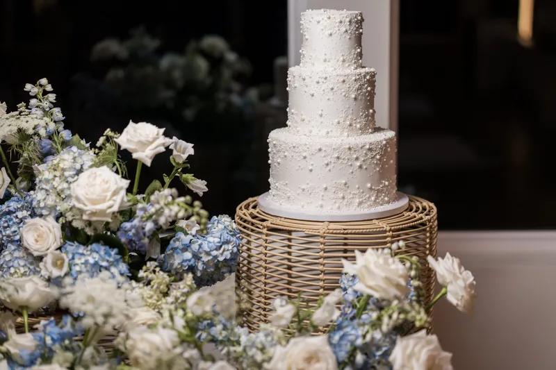 A three-tiered white textured cake sits on a woven base surrounded by blue and white flowers.