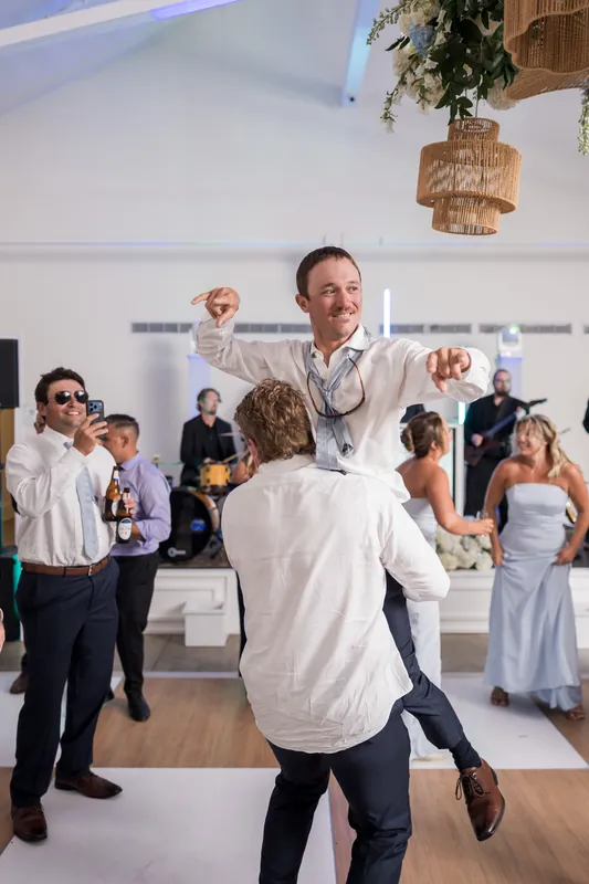 A person in a white shirt dances with arms raised as wedding guests watch from the sides of a bright indoor space.