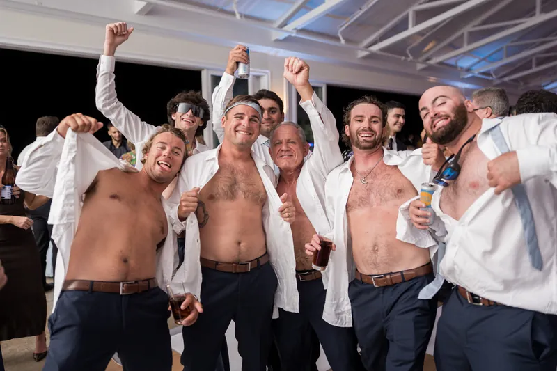 A group of shirtless men in dress pants and open white shirts raise their fists and celebrate together on the dance floor.