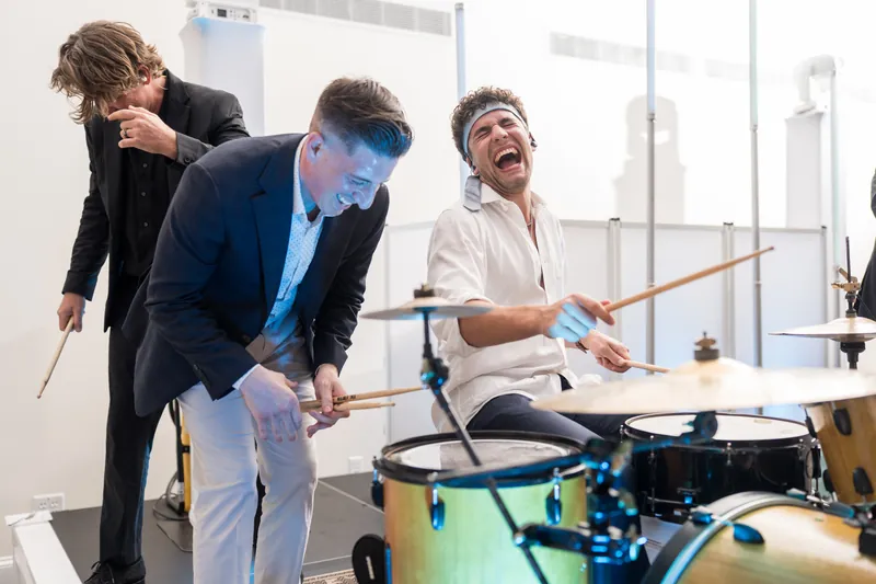 Three male guests in suits playfully engage with drums during the reception entertainment, laughing as they hold drumsticks.