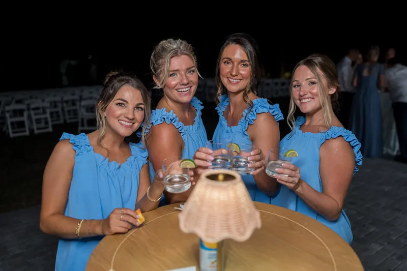 Four women in matching blue sleeveless dresses hold champagne glasses together outdoors during the reception under evening lighting.