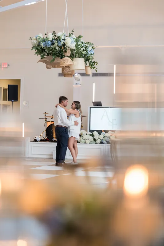 Two people in light-colored outfits embrace under a suspended floral arrangement inside the venue.