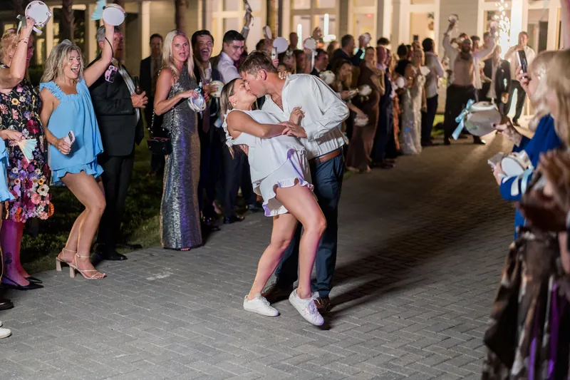 Guests line both sides of an outdoor stone pathway as two people in white and navy walk through cheering crowds.