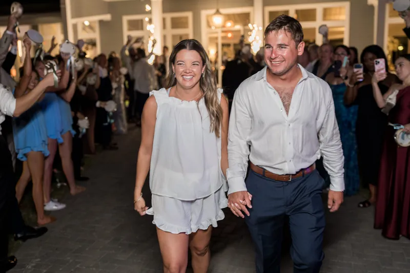 Lexie and Ed walk hand-in-hand down an outdoor corridor surrounded by applauding wedding guests.