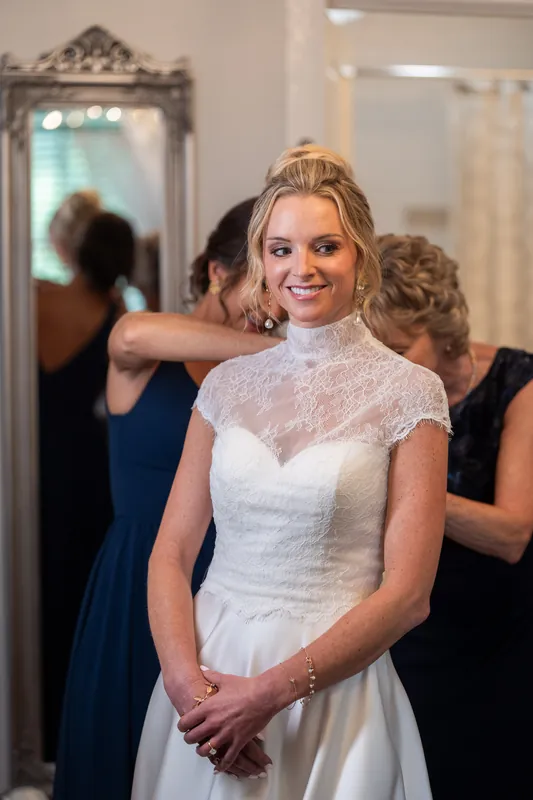 A person in a white lace wedding gown stands with hands clasped, flanked by attendants in dark clothing.