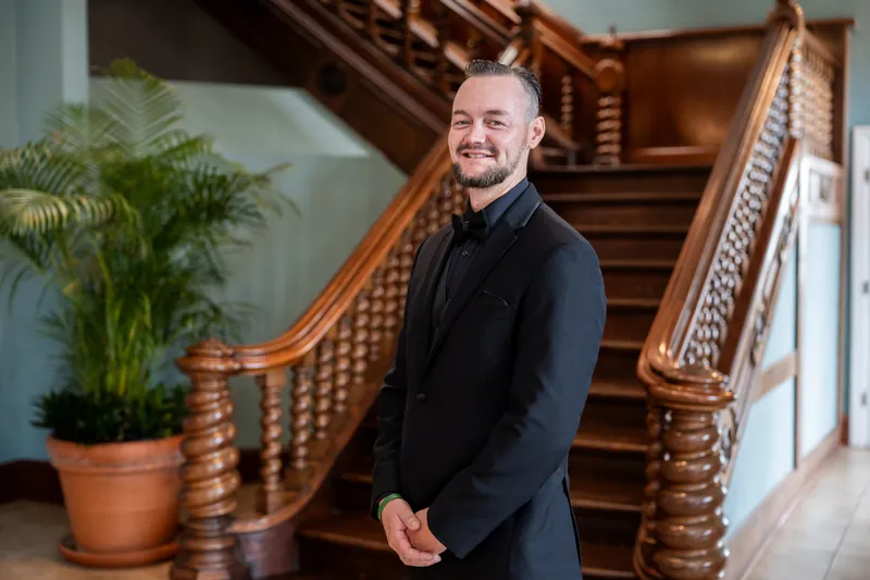 A person in a black suit smiles at the camera while standing on a wooden staircase beside a potted fern.