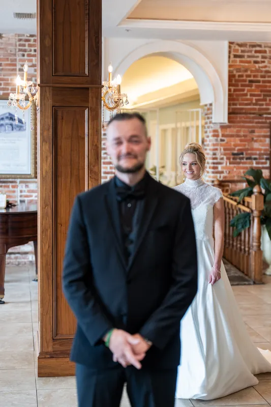 A person in a dark suit stands in an interior hallway with exposed brick, as another person in white appears behind them.