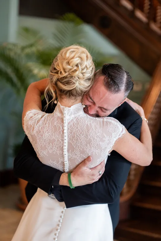 Two people embrace on a wooden staircase, one wearing a white gown and the other in a dark suit.