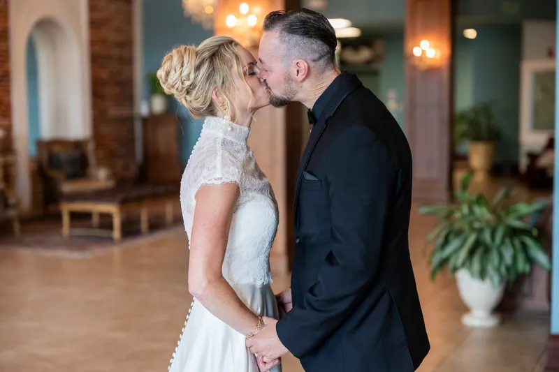 Two people kiss while standing in an elegant interior space with plants and warm lighting visible behind them.