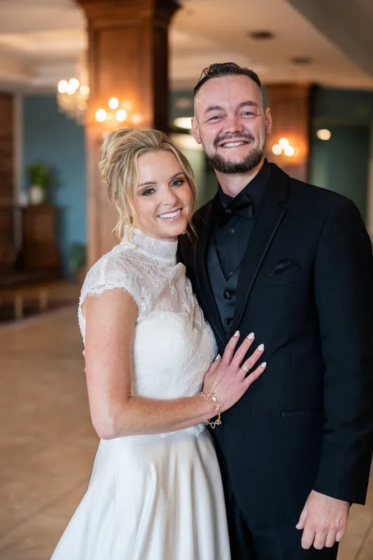 Li and Cody pose together indoors, smiling at the camera in their wedding attire under warm lighting.