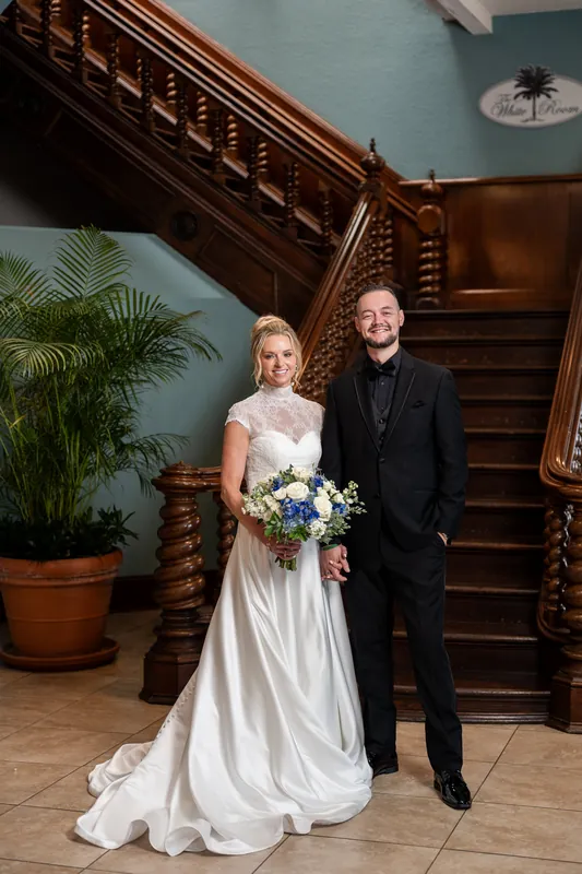 Li and Cody stand by an ornate wooden staircase, with Li holding a bouquet and Cody's arm around their shoulder.