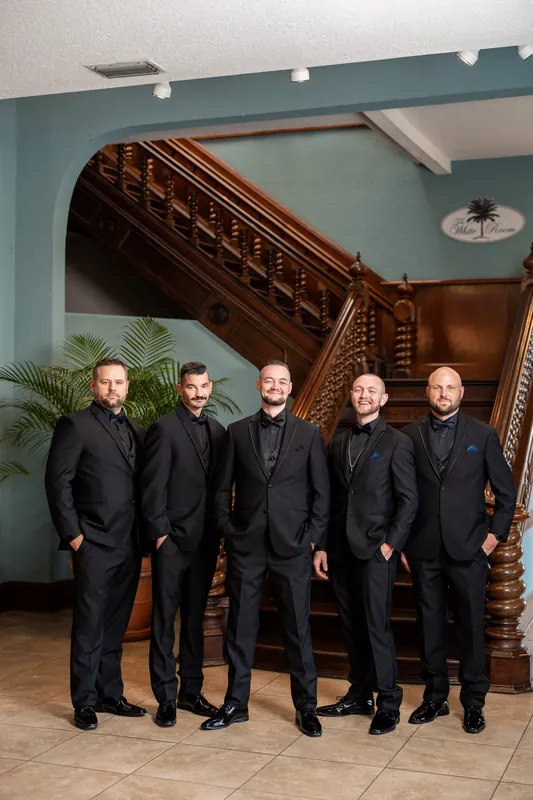 Five people in formal black suits stand together in front of a carved wooden staircase with teal walls.