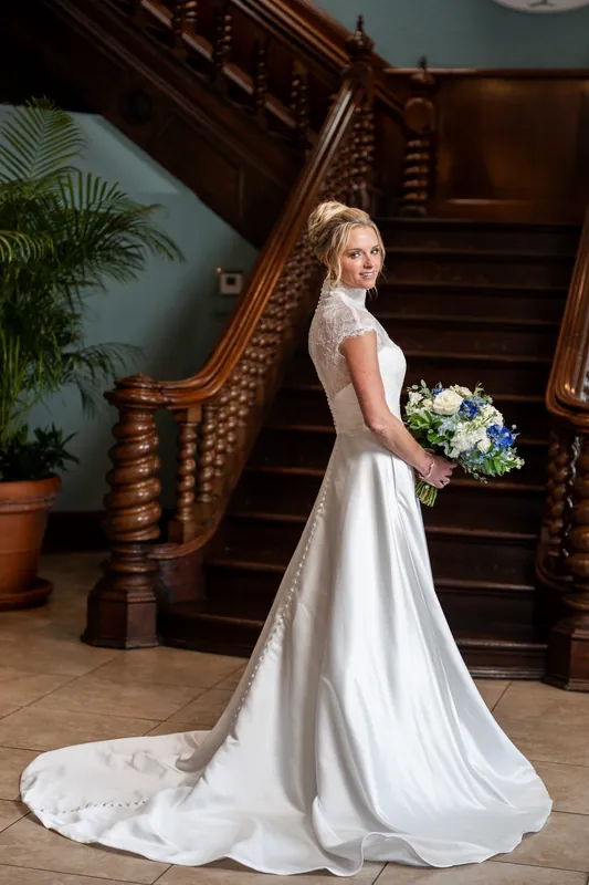 Someone in a flowing white gown with a train holds a bouquet while posing in profile near the wooden staircase.