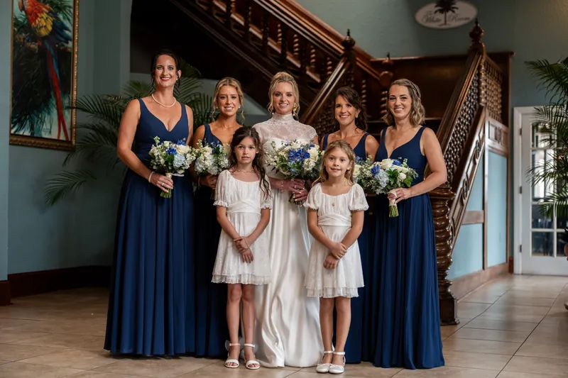 A wedding party of eight people—some in navy dresses, others in white, and one in a flowing gown—hold bouquets by the staircase.