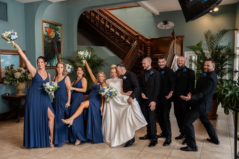 The full wedding party of fourteen people, dressed in navy, white, and black formal wear, gathers together in front of the ornate wooden staircase.